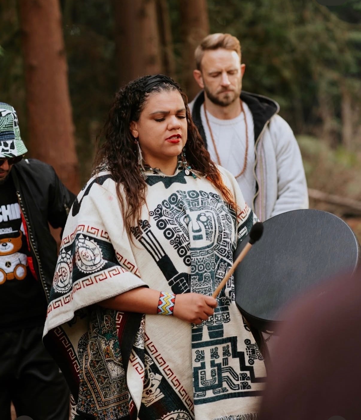 Katie holding a frame drum in a patterned poncho at a woodland cacao and hape ceremony