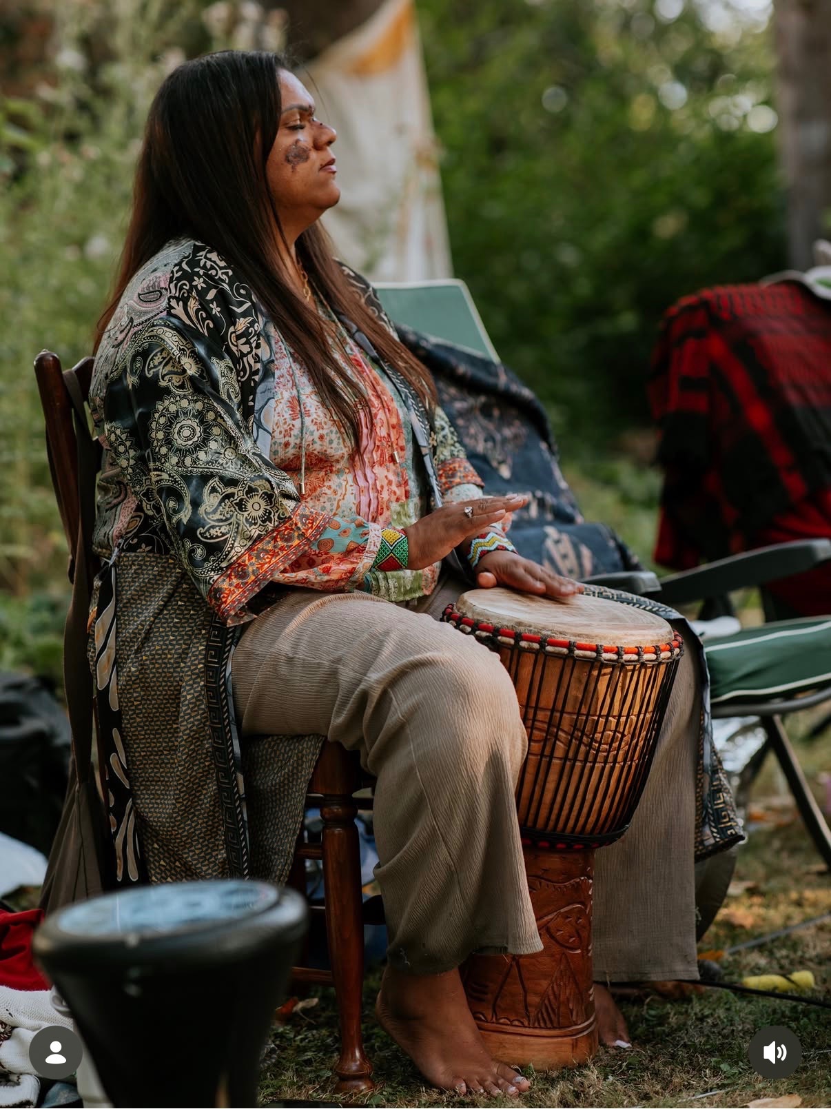A ceremony guest seated with a djembe drum, eyes closed, in a woodland clearing