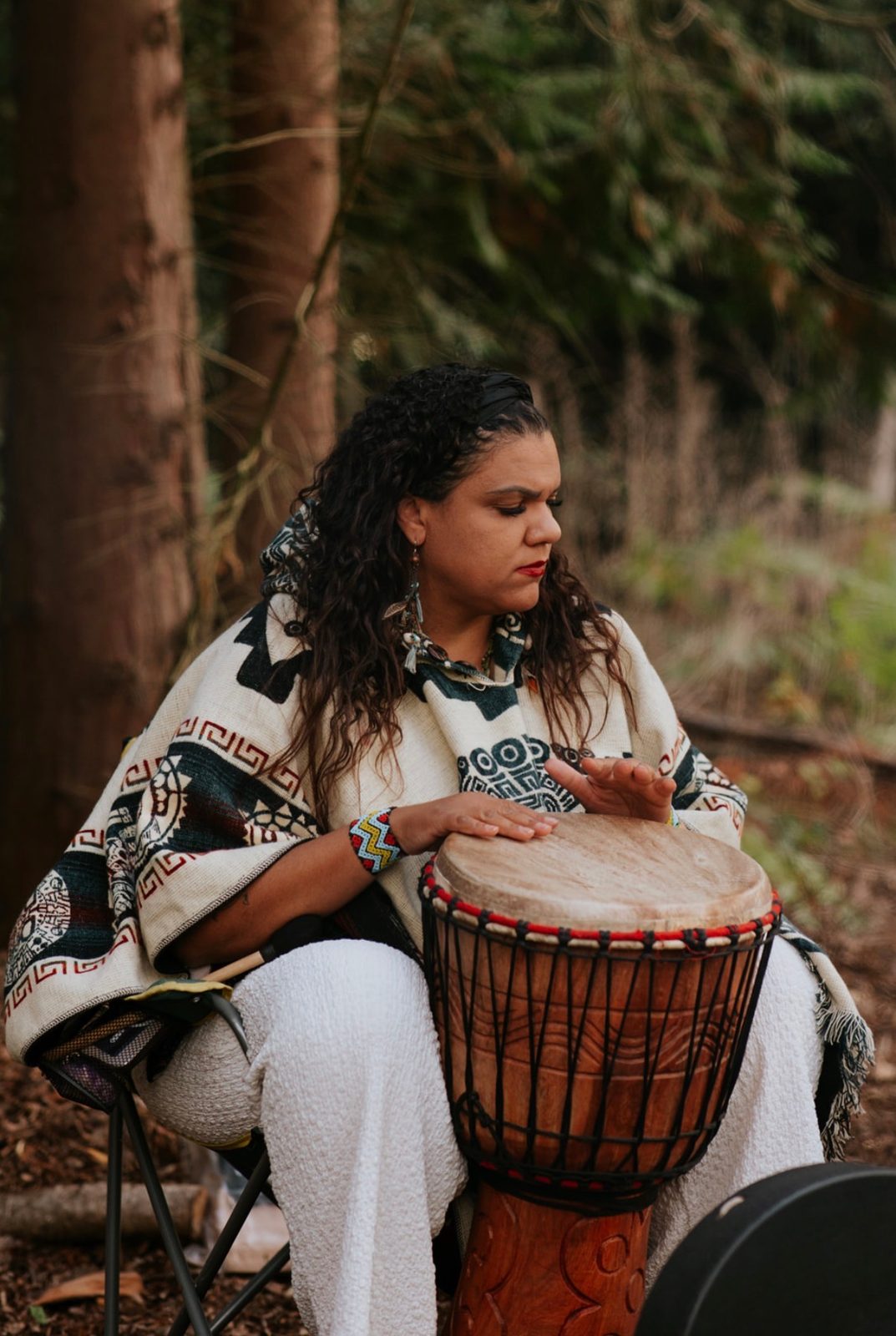 Woman playing a djembe drum in the woods, wrapped in a patterned blanket